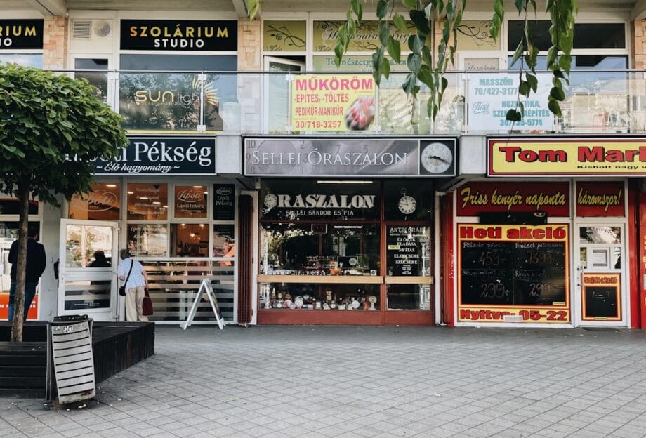 A row of storefronts with signs in Hungarian, featuring a bakery, a salon, and other shops on a pedestrian street.