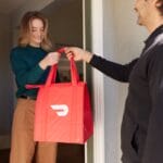A woman receives a red insulated food delivery bag from a man at the front door.