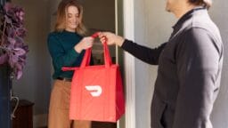 A woman receives a red insulated food delivery bag from a man at the front door.