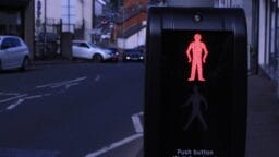 A pedestrian crossing signal displaying a red standing figure, indicating wait to cross, with a street in the background.