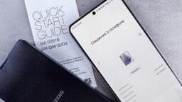 A Samsung smartphone displays device information next to a Samsung quick start guide and a black Samsung case on a gray surface.