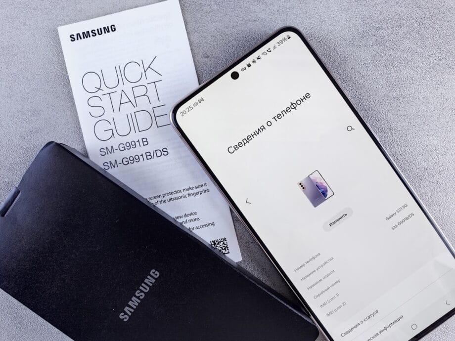Samsung smartphone displaying device information next to a quick start guide and a Samsung-branded case on a grey surface.