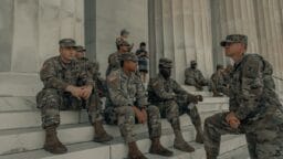 A group of soldiers in uniform sit and stand on marble steps outside a large stone building with columns.
