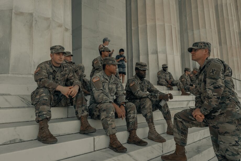 A group of soldiers in uniform sit and stand on marble steps outside a large stone building with columns.
