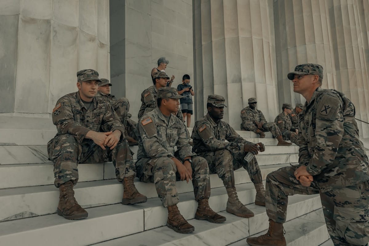 A group of soldiers in uniform sit and stand on marble steps outside a large stone building with columns.