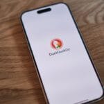 A smartphone on a wooden table displays the DuckDuckGo search engine logo and name on its screen.
