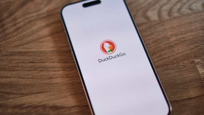 A smartphone on a wooden table displays the DuckDuckGo search engine logo and name on its screen.