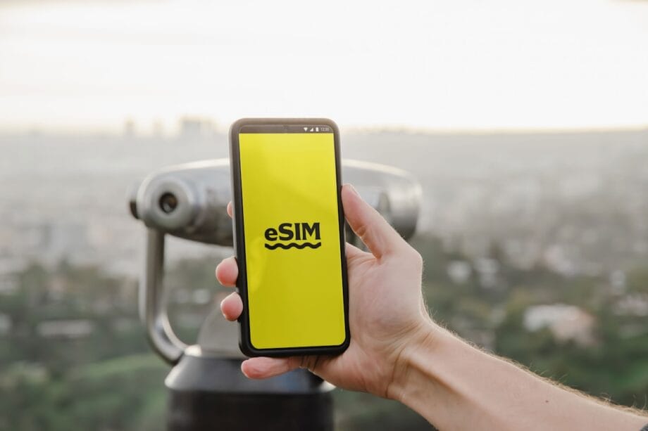 A hand holds a smartphone displaying a yellow eSIM screen in front of a scenic city view and a coin-operated telescope.