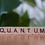 Wooden Scrabble tiles spell out the word QUANTUM on a table, with a blurred green background.