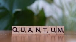 Wooden Scrabble tiles spell out the word QUANTUM on a table, with a blurred green background.