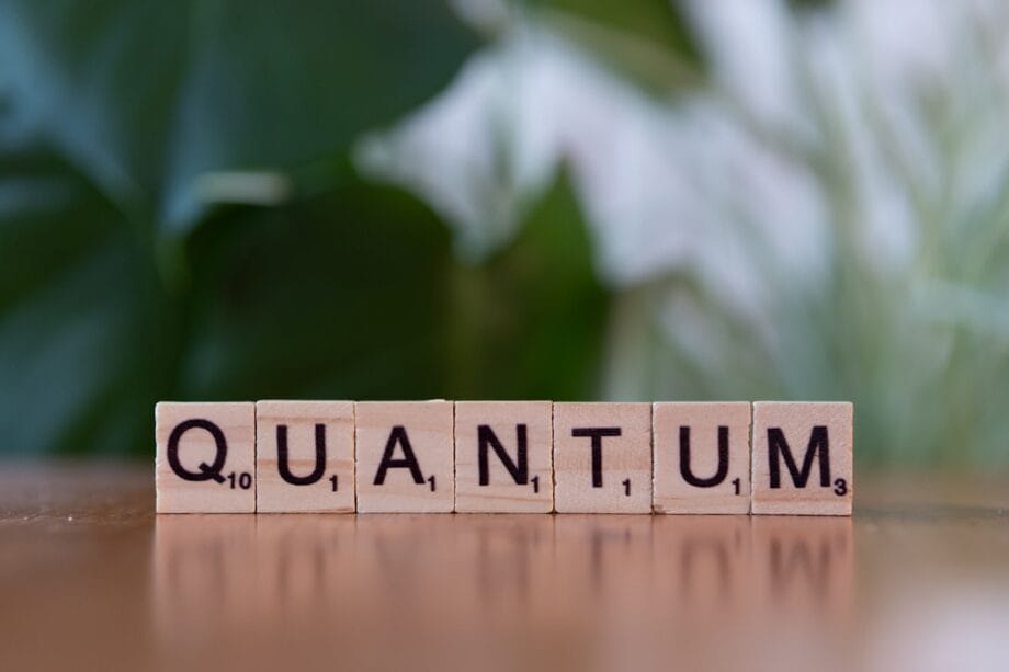 Wooden Scrabble tiles spell out the word QUANTUM on a table, with a blurred green background.