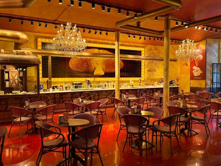 A brightly lit café with red chairs and tables, chandeliers, and a counter in front of a large mural of food on the wall.