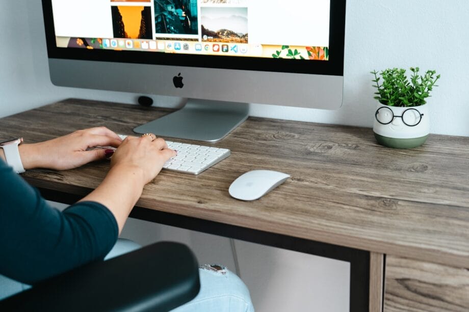 Person typing on a keyboard at a wooden desk with an iMac, mouse, and a small potted plant beside the monitor.