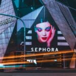 Sephora store at night with a large illuminated makeup advertisement on the geometric glass facade and light trails from passing cars.