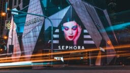 Sephora store at night with a large illuminated makeup advertisement on the geometric glass facade and light trails from passing cars.