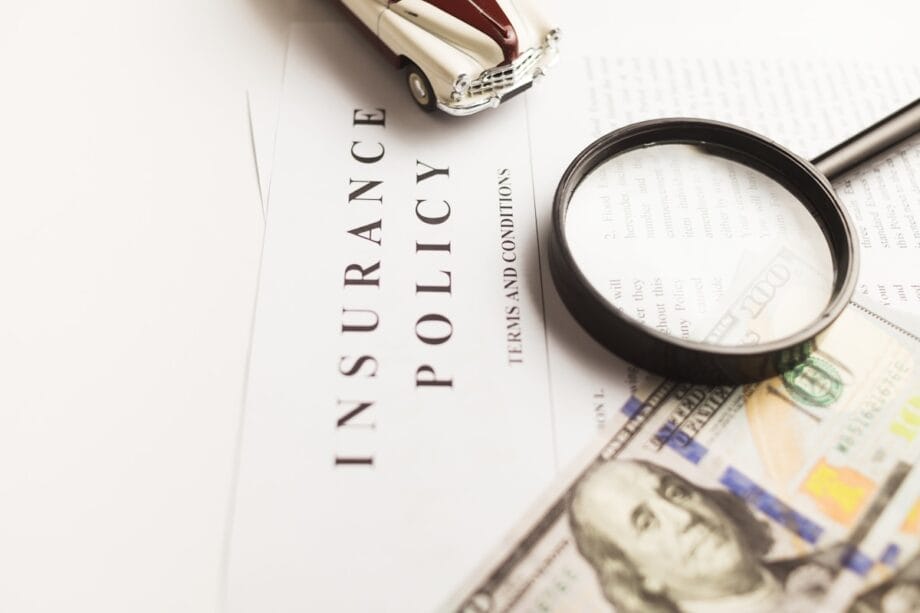 A magnifying glass, toy car, and a 100 bill on top of an insurance policy document.