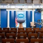 Empty White House press briefing room with podium, American flag, blue backdrop, rows of chairs, and cameras set up on the side.