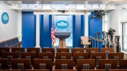 Empty White House press briefing room with podium, American flag, blue backdrop, rows of chairs, and cameras set up on the side.