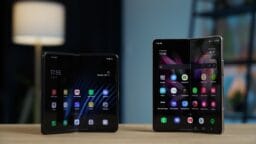 Two foldable smartphones are displayed open on a wooden table, each showing their home screens with various app icons.