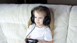 A young girl wearing headphones sits on a couch holding a video game controller.