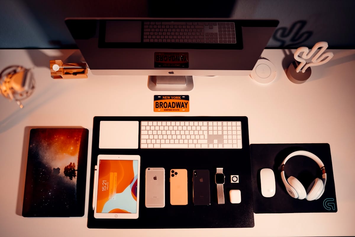 Neatly organized desk with an iMac, keyboard, iPad, various smartphones, AirPods, headphones, mouse, and a Broadway souvenir.
