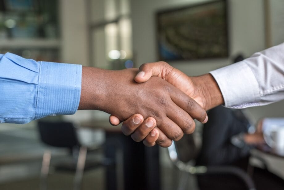 Two people in dress shirts shaking hands in an office setting, with blurred desks and chairs in the background.