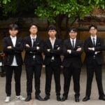 Five young men in black suits and ties stand side by side outdoors with arms crossed, facing the camera.