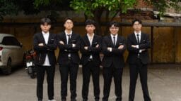 Five young men in black suits and ties stand side by side outdoors with arms crossed, facing the camera.