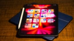 A tablet with a stylus and case displays various app icons on a wooden surface, illuminated with a red and black background.