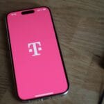 A smartphone displaying the T-Mobile logo on a pink screen rests on a wooden surface next to a black notebook.