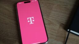A smartphone displaying the T-Mobile logo on a pink screen rests on a wooden surface next to a black notebook.