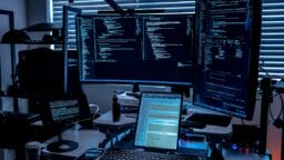 A workspace with multiple monitors displaying code, a keyboard, coffee cup, and tech devices, set against window blinds.