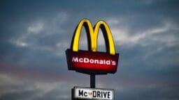 Illuminated McDonalds sign with the golden arches and McDrive sign underneath against a cloudy evening sky.