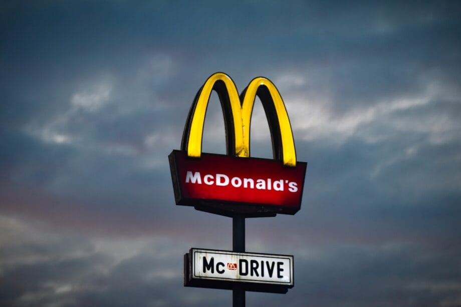 Illuminated McDonalds sign with the golden arches and McDrive sign underneath against a cloudy evening sky.