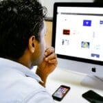 A person sits at a desk, looking at a computer screen displaying image thumbnails, with a phone and wallet nearby.