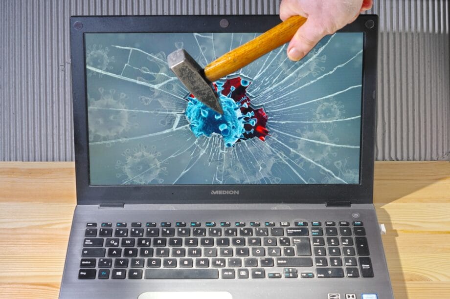 A hand with a hammer smashes a laptop screen displaying a virus image, causing the screen to crack.