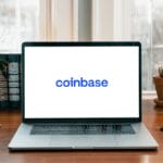 A laptop on a wooden desk displays the Coinbase logo on its screen, with plants and books in the background.