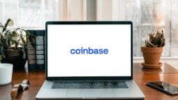 A laptop on a wooden desk displays the Coinbase logo on its screen, with plants and books in the background.