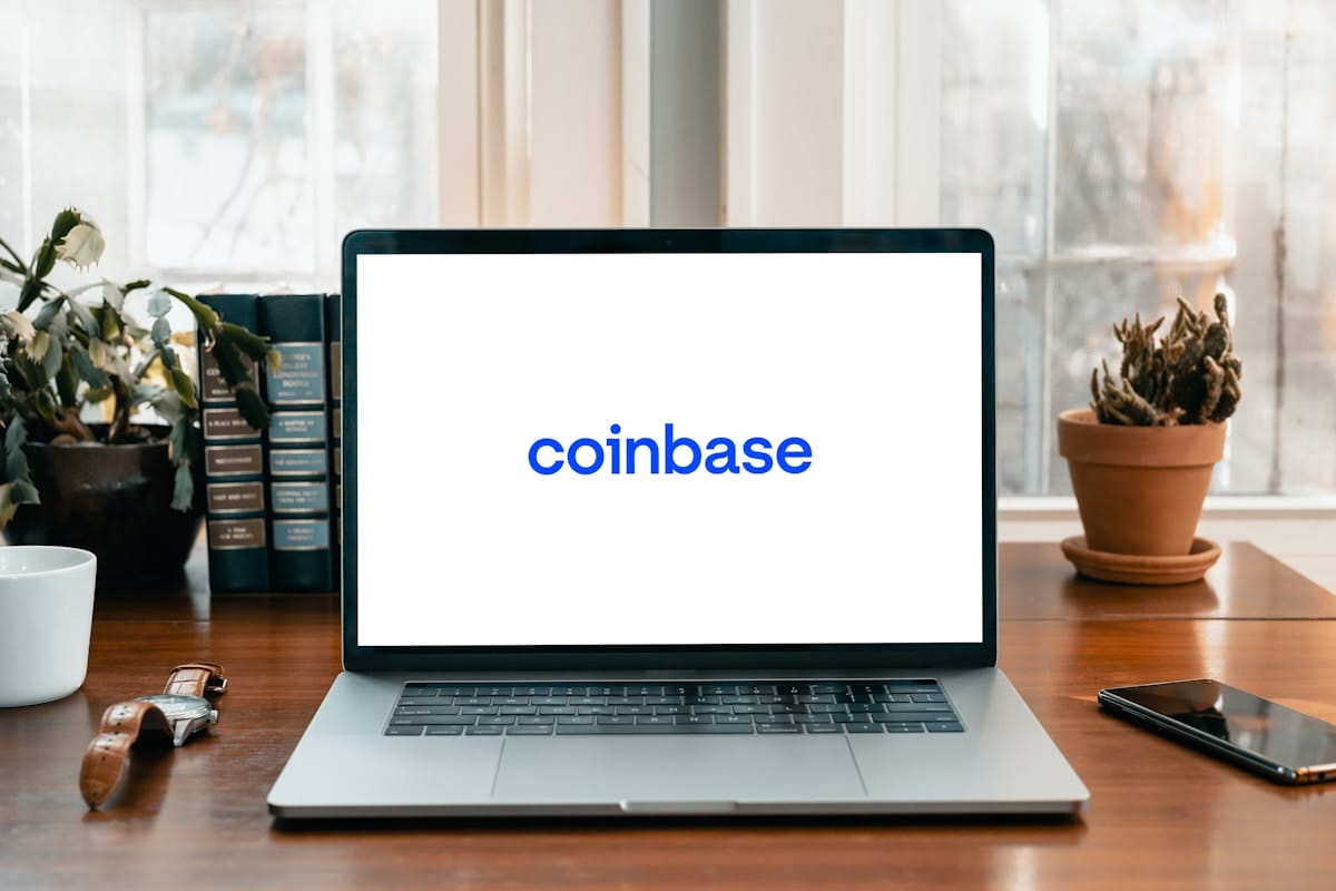 A laptop on a wooden desk displays the Coinbase logo on its screen, with plants and books in the background.