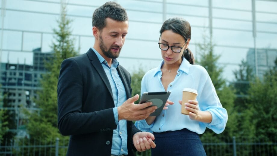 Two people standing outdoors looking at a tablet; one holds a coffee cup. A glass building and trees are in the background.