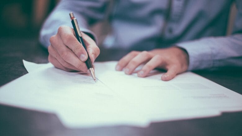 Person in a blue shirt writes on paper documents with a pen at a desk.