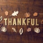 Gold glitter letters spelling THANKFUL surrounded by wooden leaves, pumpkins, and acorns on a brown wooden surface.