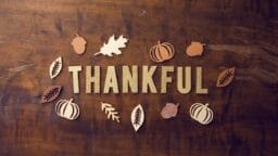 Gold glitter letters spelling THANKFUL surrounded by wooden leaves, pumpkins, and acorns on a brown wooden surface.