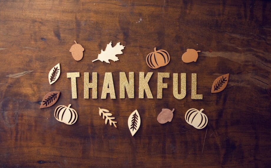 Gold glitter letters spelling THANKFUL surrounded by wooden leaves, pumpkins, and acorns on a brown wooden surface.