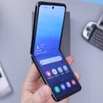 A hand holds a modern foldable smartphone, with two older cell phones and a plant visible on the desk in the background.