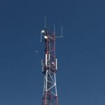 A red and white telecommunications tower stands against a clear blue sky with the moon visible in the background.