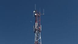A red and white telecommunications tower stands against a clear blue sky with the moon visible in the background.