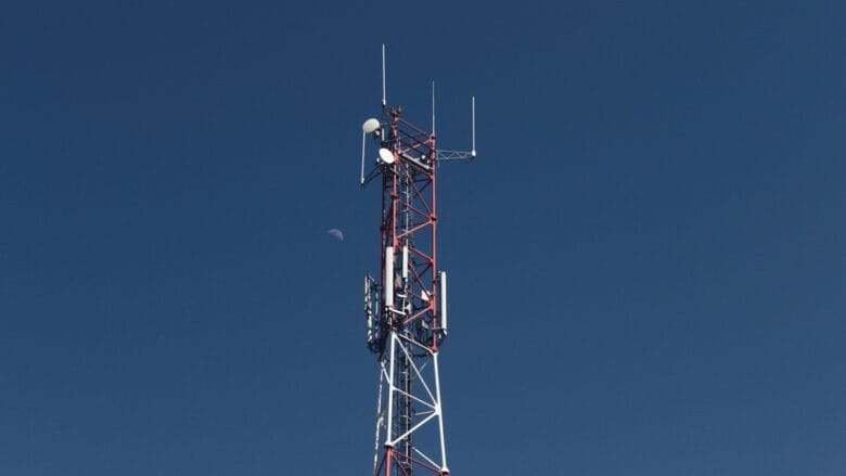 A red and white telecommunications tower stands against a clear blue sky with the moon visible in the background.