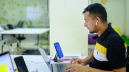 A man wearing wireless earbuds works on a laptop at a desk in a modern, brightly lit office space.