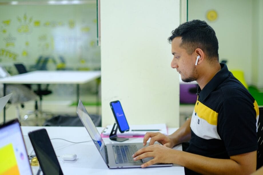 A man wearing wireless earbuds works on a laptop at a desk in a modern, brightly lit office space.